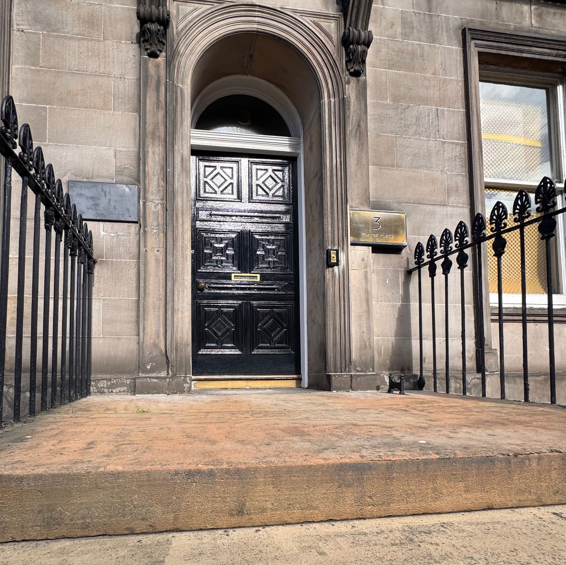 The main door to the practice, a black door with a brass letterbox and plaque, framed by its carved stone archway and iron railings