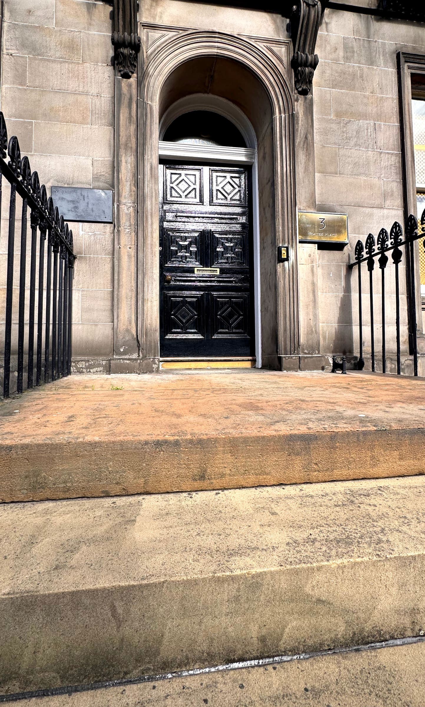 The black front door of 3 Coates Place, framed by its carved stone archway, brass plaque and iron railings
